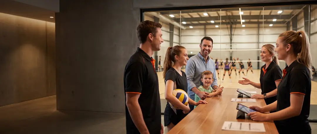 Front-desk staff checking in players and families at a busy sports facility, with a volleyball court behind the glass